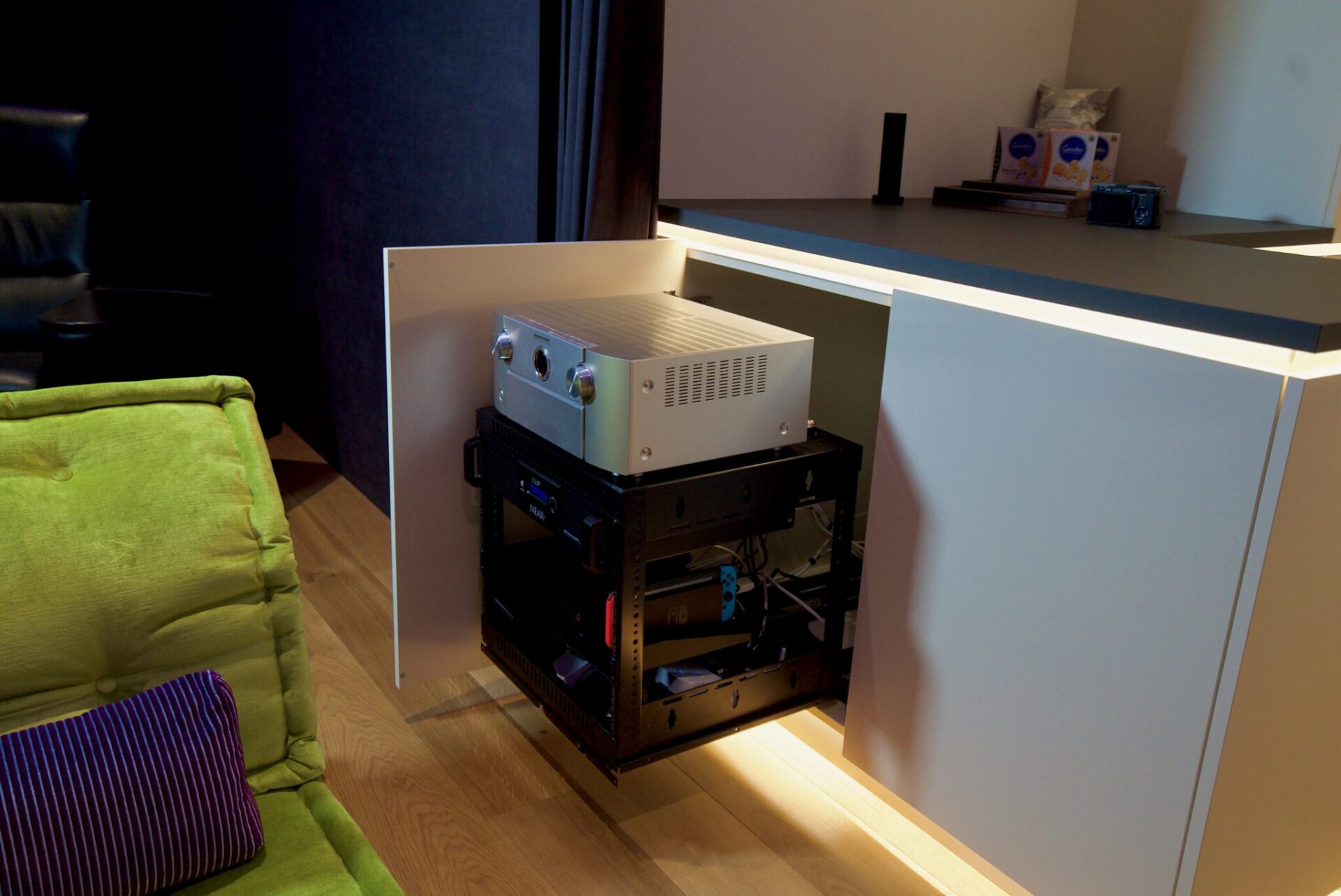 Nicht nur Süßigkeiten haben ihren Platz in der Küchenzeile. Auch die Technik versteckt sich hier. Dank eines professionellen Technik-Racks ist alles sauber und sicher verstaut, kann aber bei Bedarf rausgezogen werden. Wir sehen hier den Marantz SR7015 ganz oben auf dem Technik Rack. Darunter können wir noch eine extra Endstufe für die Subwoofer erkennen. Auch eine Nintendo Switch wurde hier verbaut.