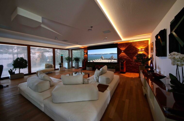 Großes Wohnraumkino mit dunklem Holzboden und weißer Couch. Der Beamer ist in die Decke integriert und über Deckenlautsprecher wird 3D Ton erzeugt.