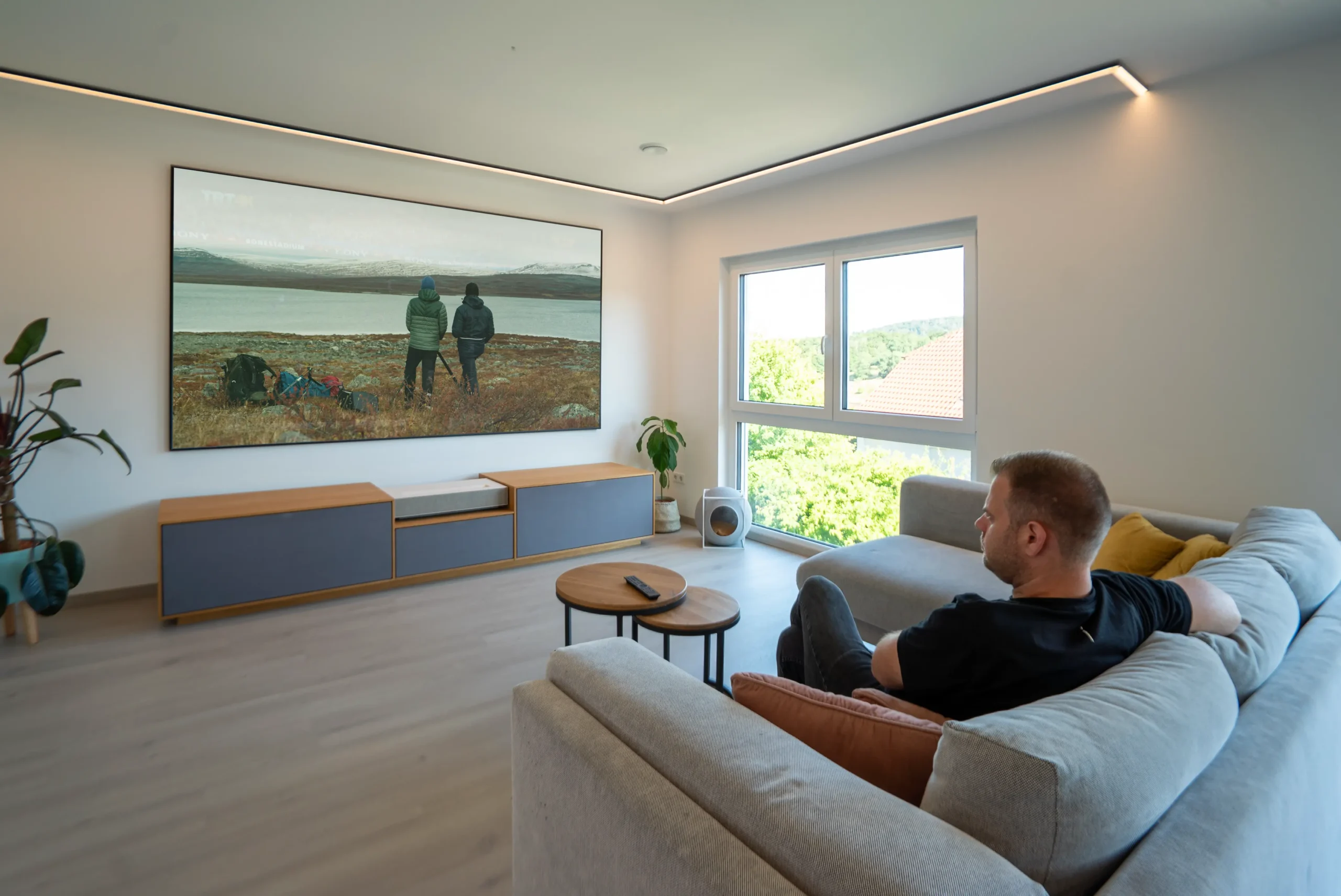 Wohnzimmer-Szene: Mann entspannt auf grauem Sofa, blickt auf 120″ ALR-Leinwand über AVITECT White-Cinema Lowboard mit geschlossenem Eiche-/Akustikstoff-Finish