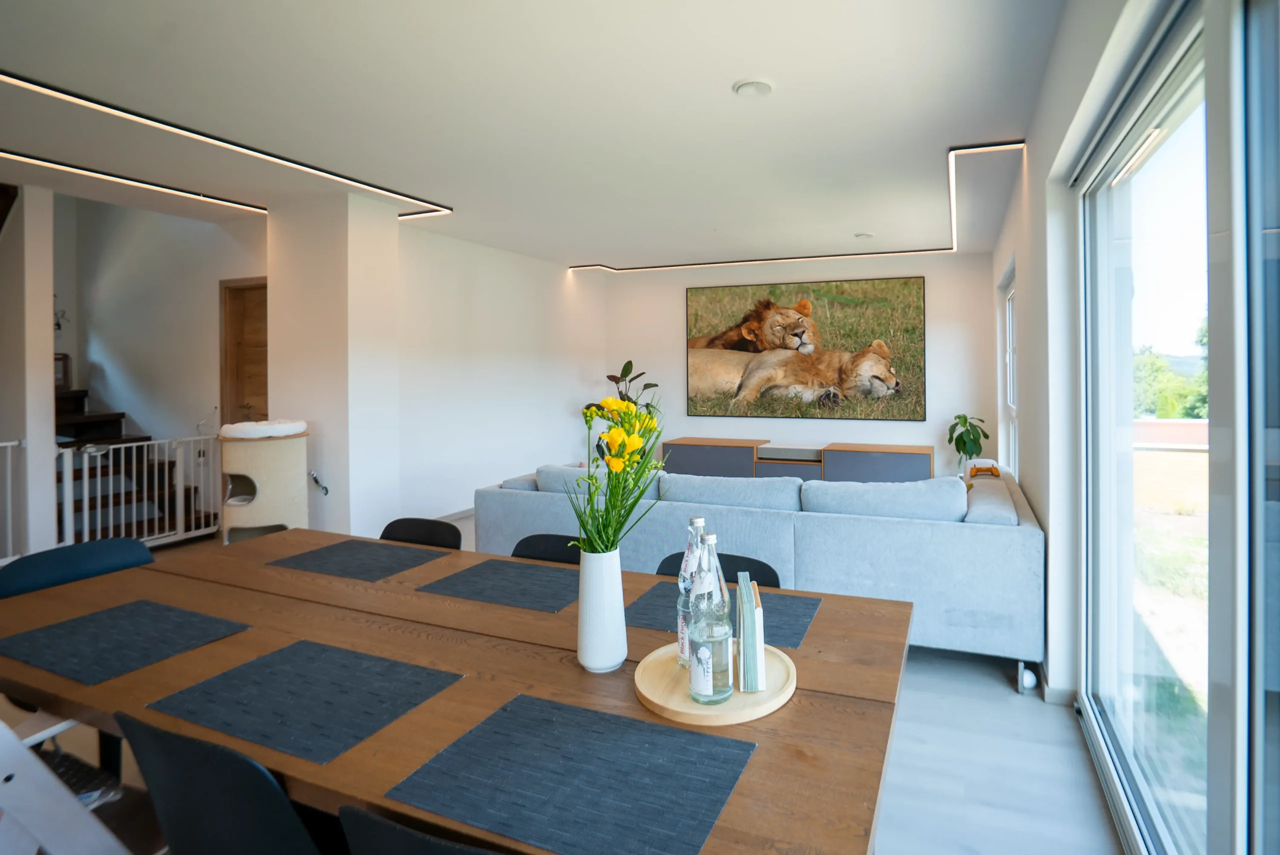 Essbereich-Perspektive: Holzesstisch mit Blumen­vase, Blick über graues Sofa auf AVITECT White-Cinema mit 120″ ALR-Leinwand und Design-Lowboard