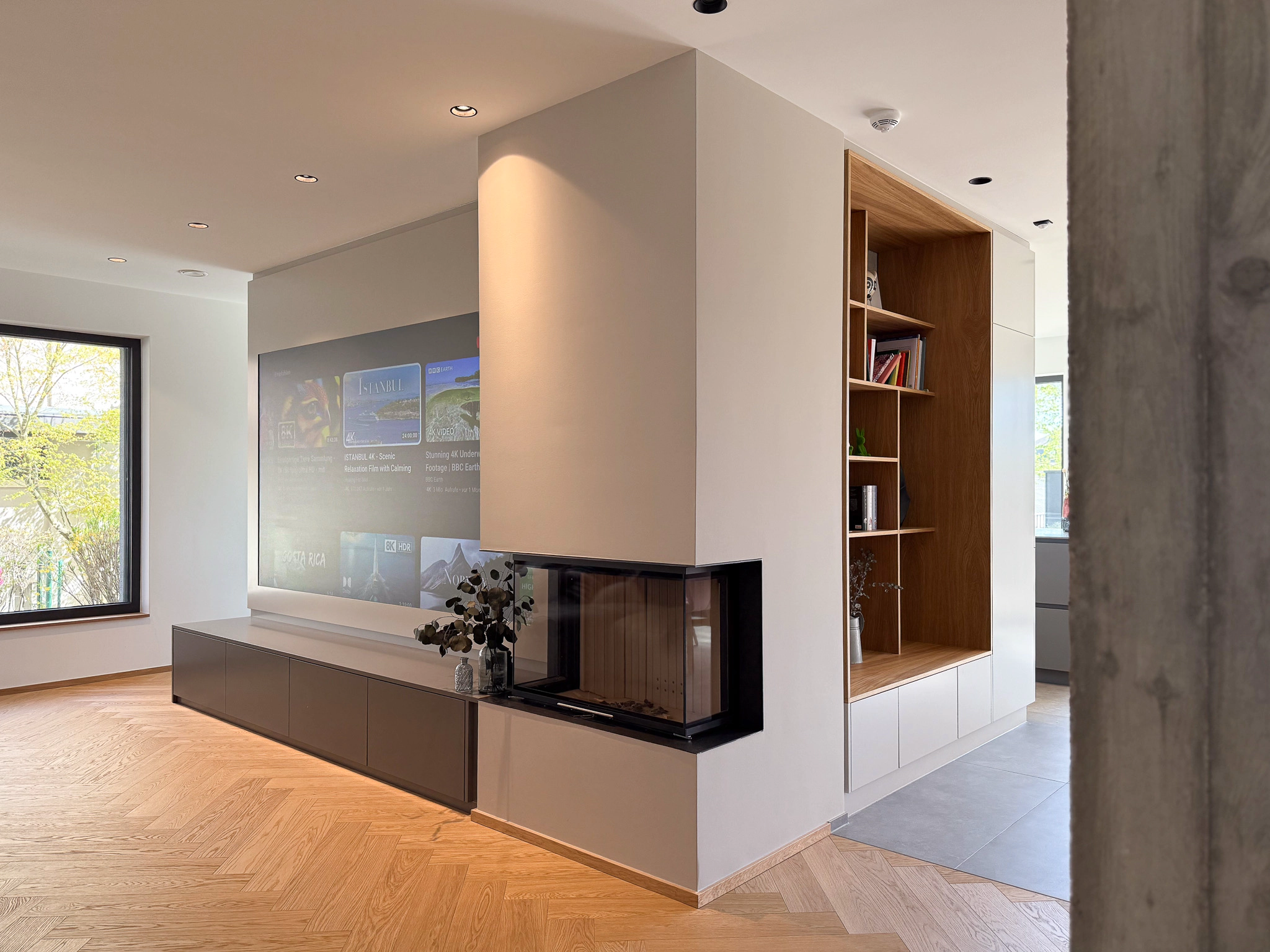 Modernes Wohnzimmer mit bündig in die Wand eingelassener Projektionsleinwand über einem schwebenden Sideboard, verglastem Eckkamin im zentralen Raumteiler und offenem Holzregal; heller Fischgrätparkettboden und großes Fenster sorgen für viel Tageslicht.