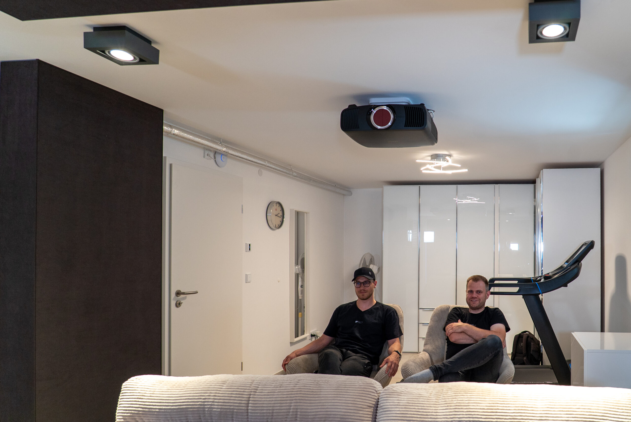 Blick in die Rückseite des Heimkinos mit Beamer an der Decke, zwei Sitzen und Laufband – multifunktionaler Raum, realisiert vom AVITECT Studio Rhein-Main.