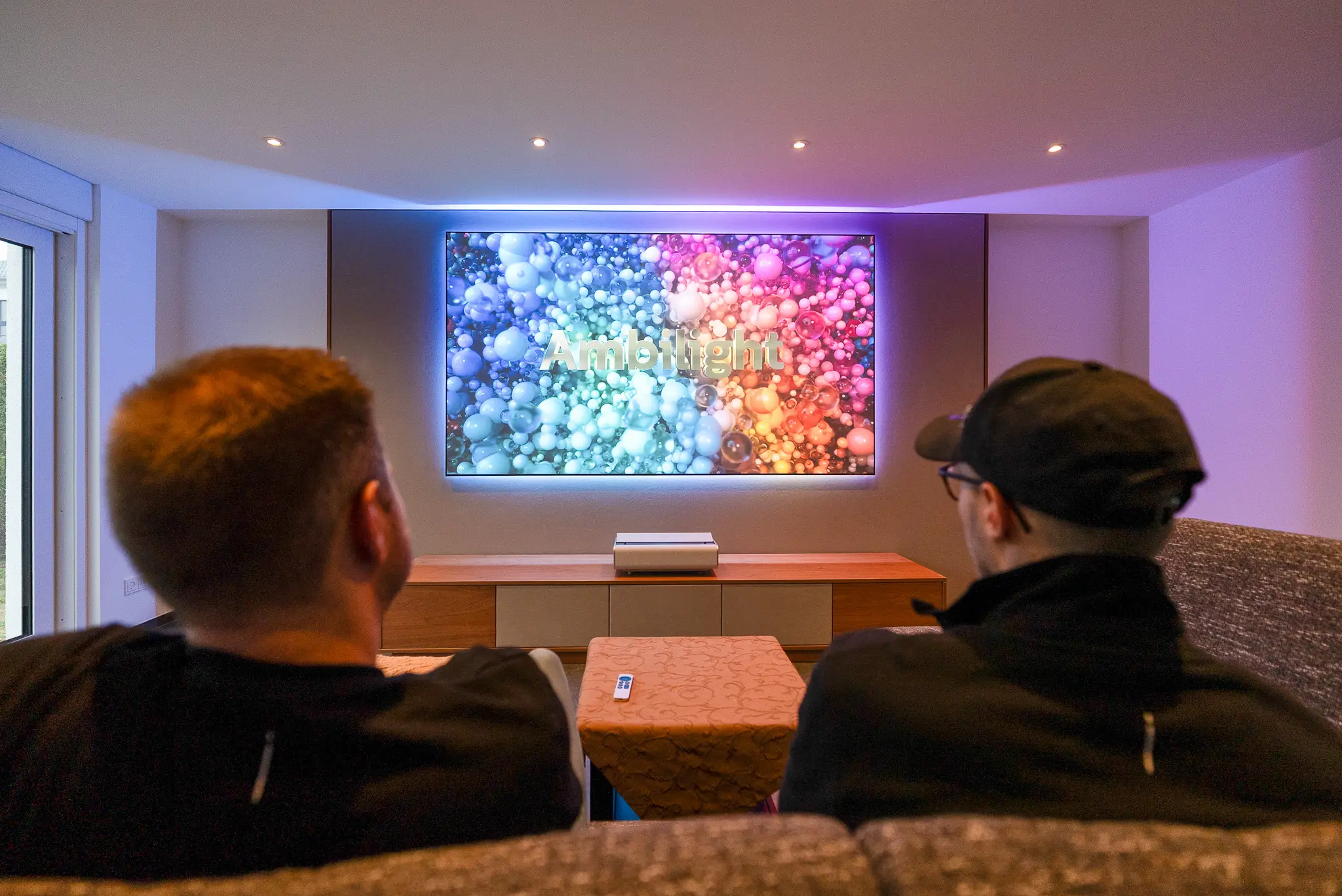 Zwei Zuschauer bestaunen den "Ambilight"-Schriftzug auf der Leinwand, dessen Farben durch das LED-System dynamisch auf die Rückwand erweitert werden.